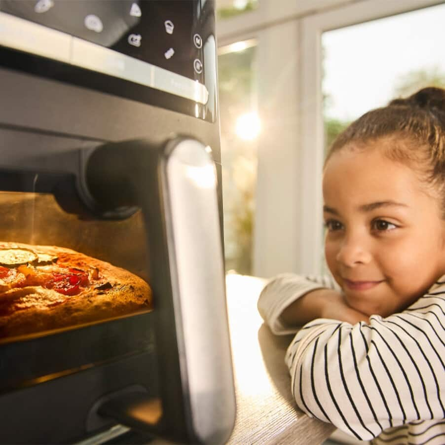 Una bambina guarda sorridente una pizza che cuoce in una friggitrice ad aria.