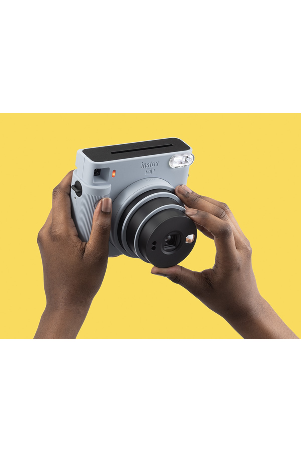 Sarı arka plan önünde gri renkli Instax SQ1 anlık kamerayı tutan bir kişi.