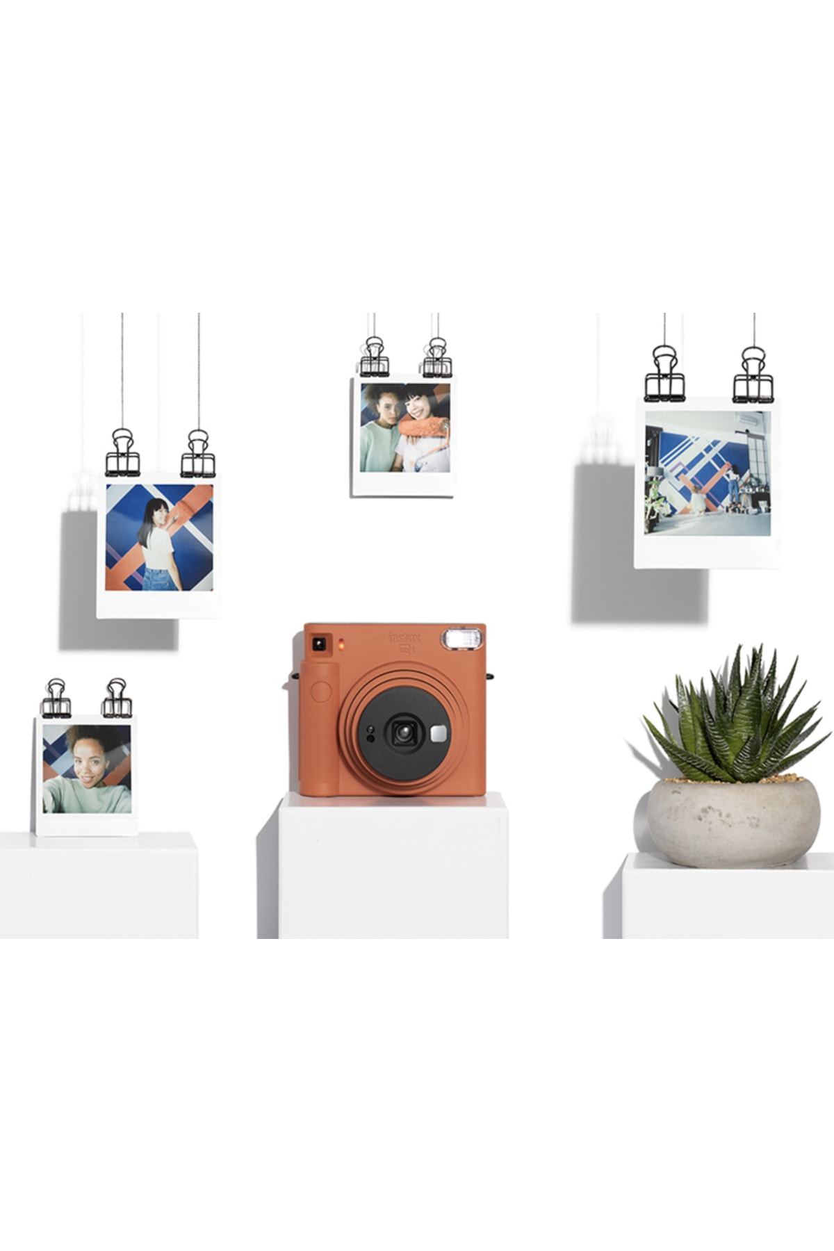 Beyaz bir kaide üzerinde turuncu bir anlık kamera, asılı polaroid fotoğraflar ve bir sukulent ile çevrili.