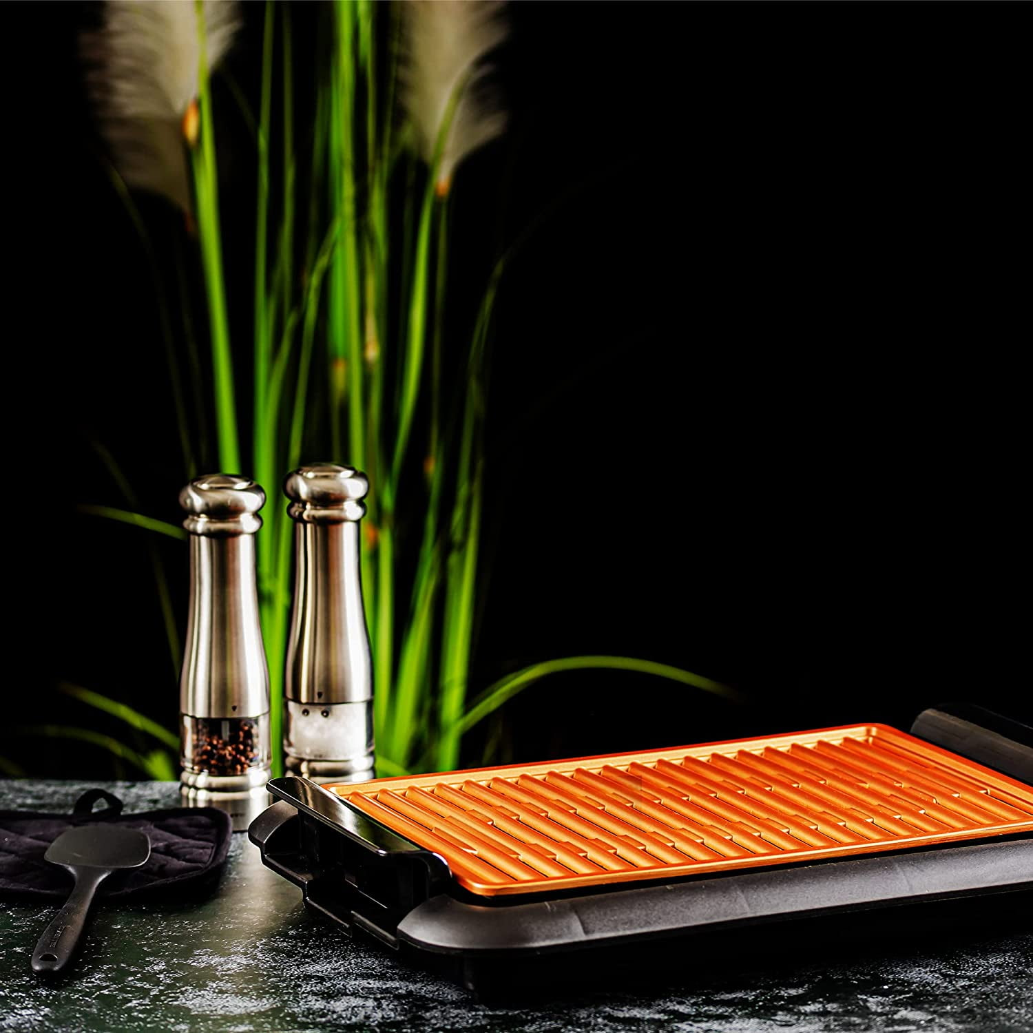 Elektrischer Grill mit orangefarbener Platte, Salz- und Pfefferstreuer sowie grüner Pflanze auf dunklem Tisch.