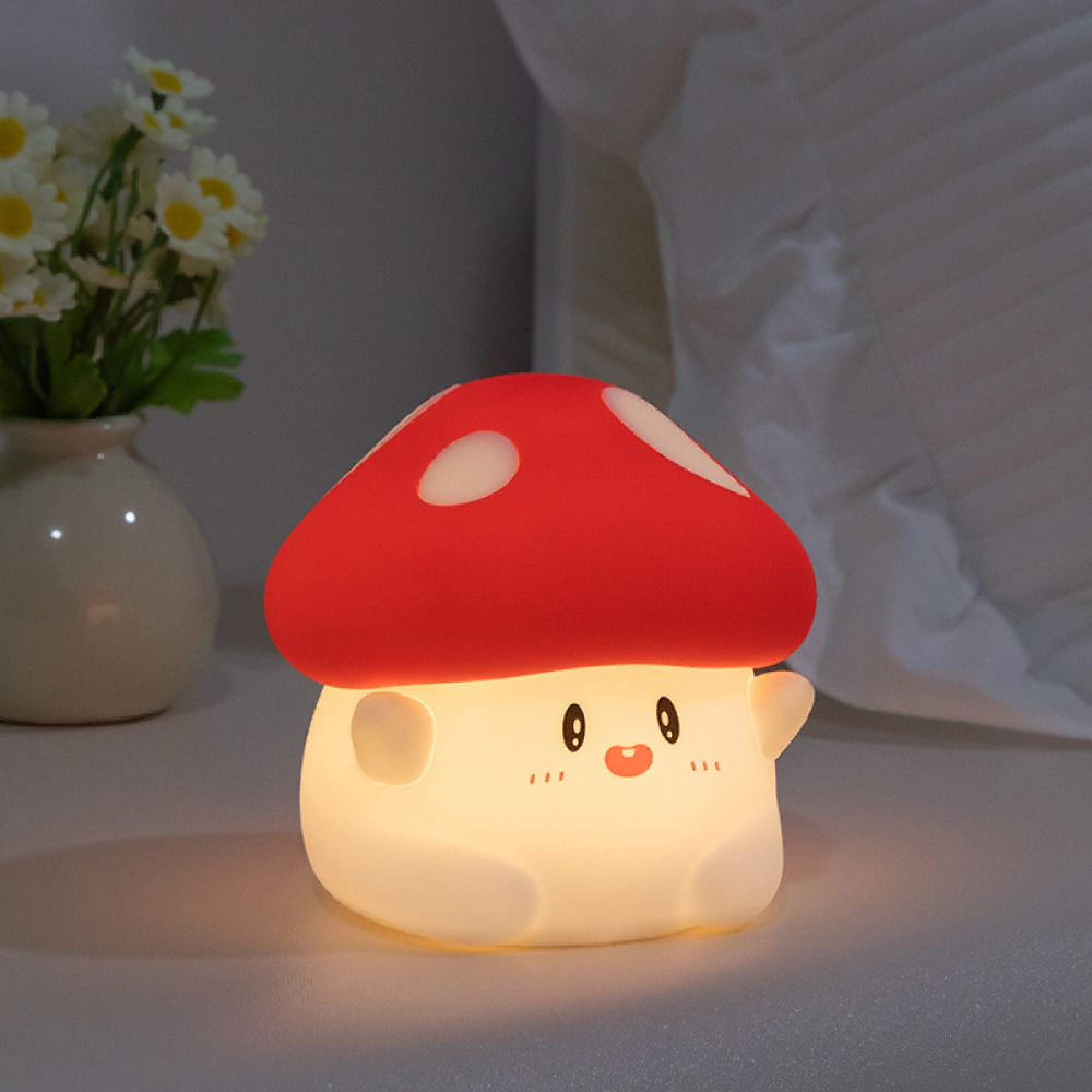 Niedliche, leuchtende Pilz-Nachtlampe mit Gesicht auf hellem Untergrund, daneben eine Vase mit weißen Blumen.