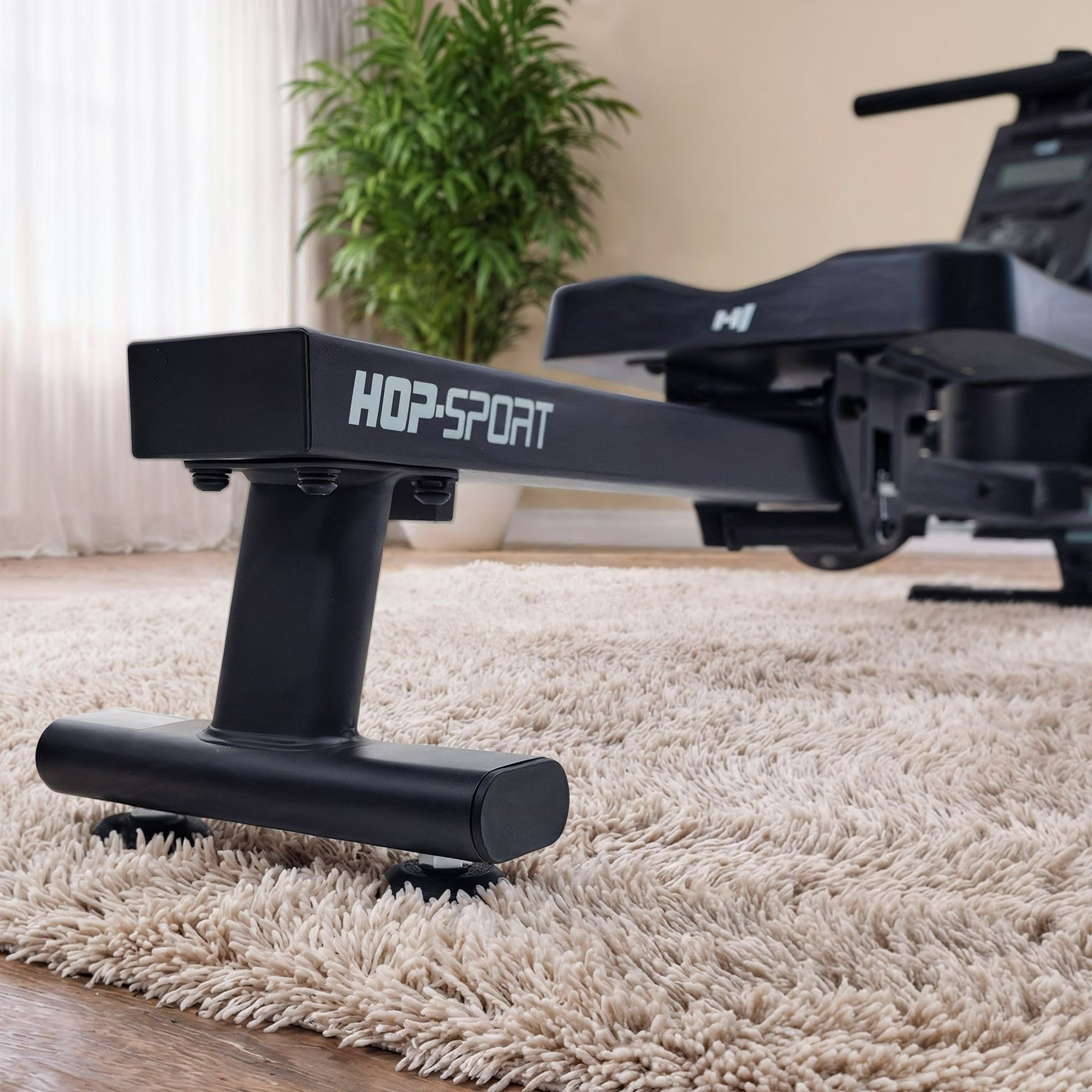 Ein schwarzes Hop-Sport Rudergerät steht auf einem flauschigen Teppich vor einer Wand und einer grünen Pflanze.