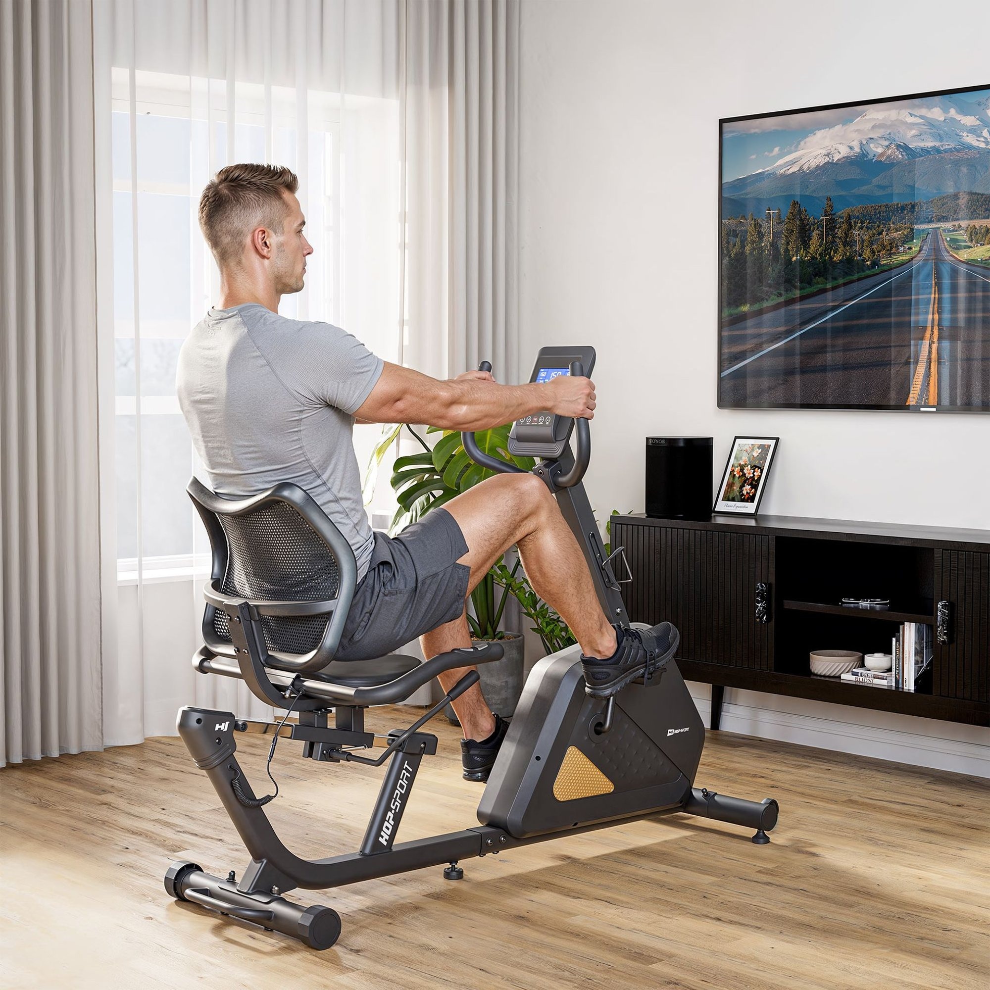 Ein Mann in Sportkleidung trainiert zu Hause auf einem Liegeheimtrainer vor einem Fernseher.