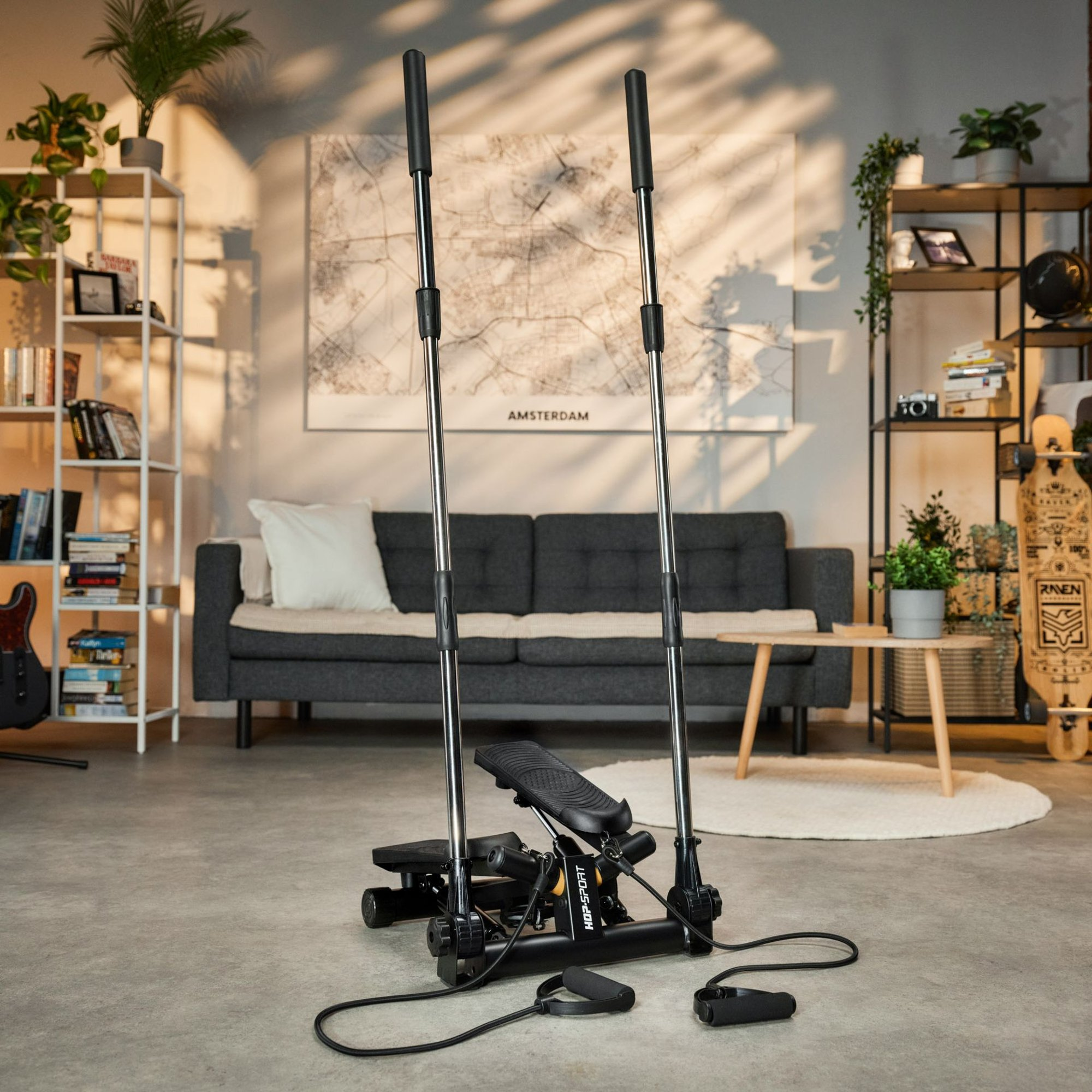 Ein schwarzer Fitness-Stepper mit langen Griffen und Widerstandsbändern in einem hellen Wohnzimmer.