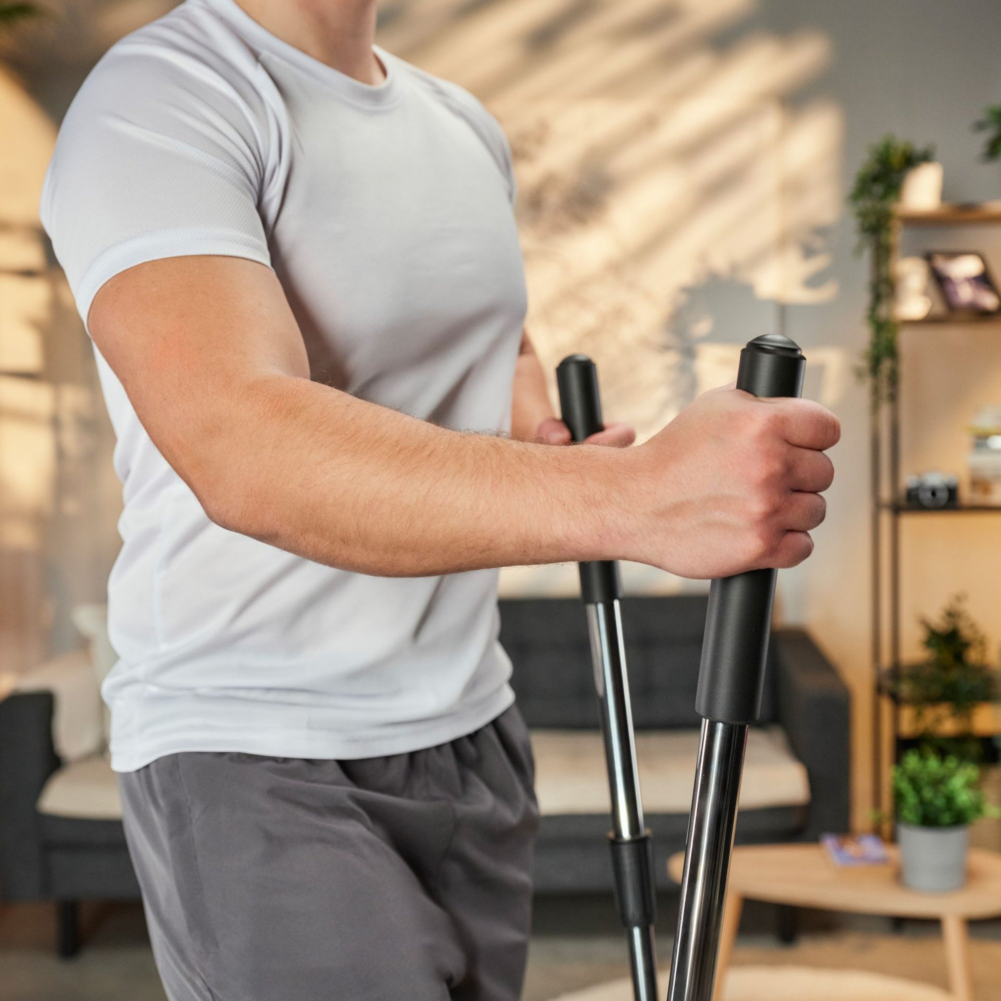 Ein Mann im weißen T-Shirt und grauen Shorts trainiert im Wohnzimmer an einem Crosstrainer.
