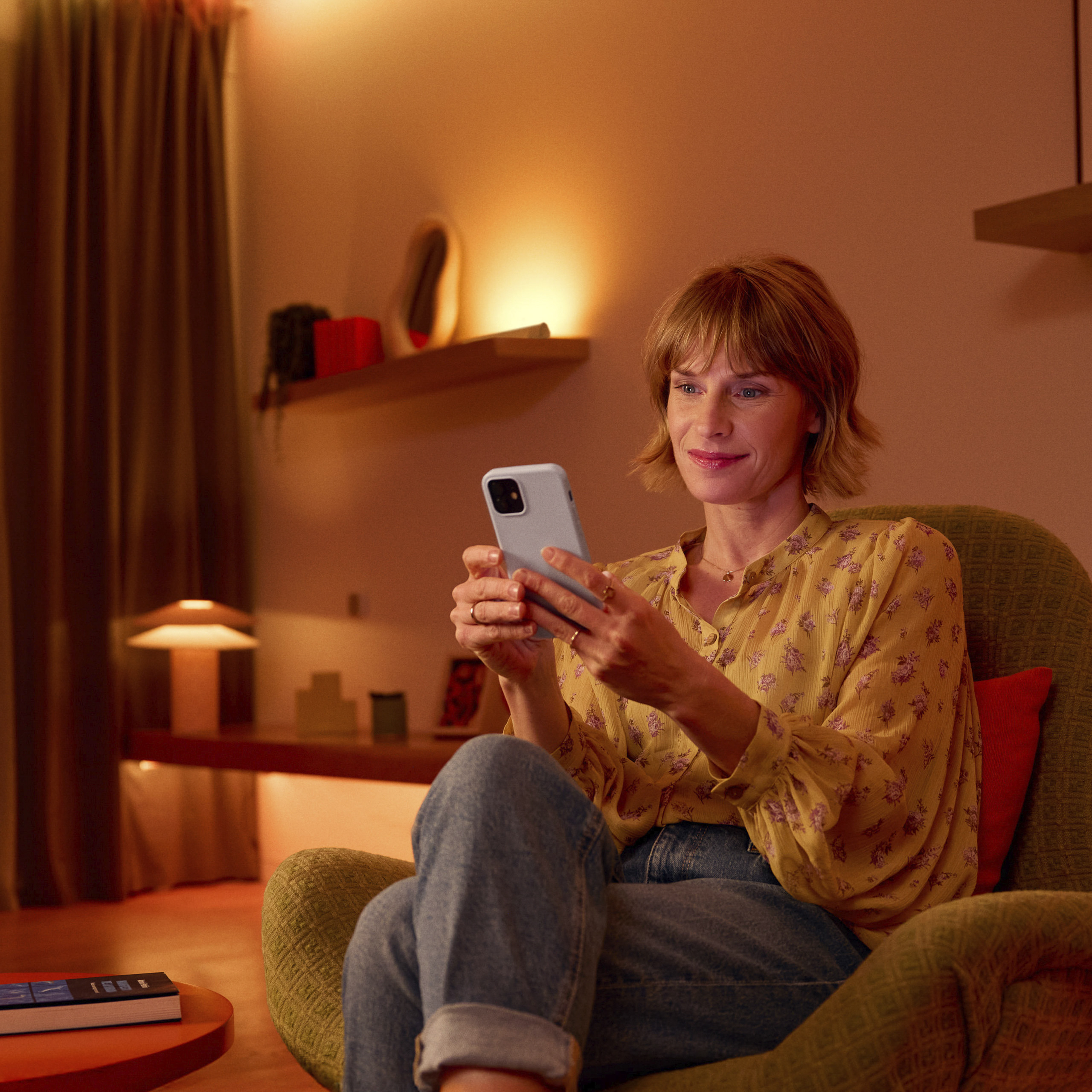 Een blonde vrouw zit in een groene fauteuil te glimlachen naar haar smartphone in een warm verlichte kamer.