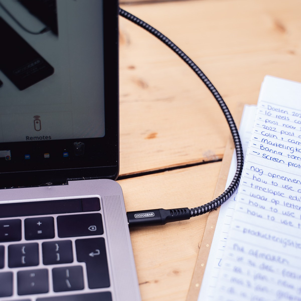 Un câble tressé noir et gris est branché sur un ordinateur portable, à côté d'un bloc-notes sur une table en bois.