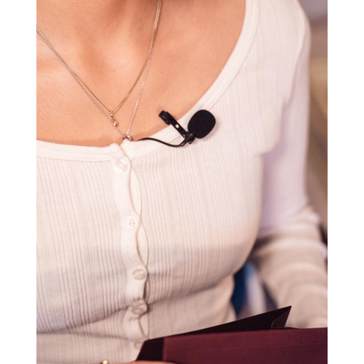 A woman wearing a white ribbed top has a small lavalier microphone clipped to her blouse.