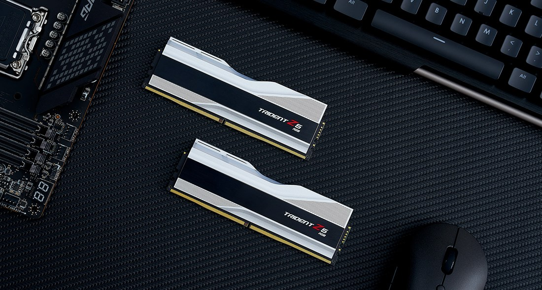 Dos módulos de RAM Trident Z5 RGB plateados y negros sobre una superficie negra texturizada junto a placa base, teclado y ratón.