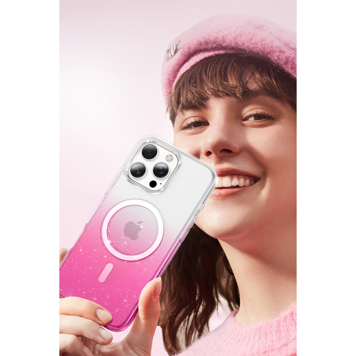 Pembe kıyafetli gülen bir kadın, pembe arka planda pembe-beyaz MagSafe kılıflı parıltılı bir iPhone tutuyor.