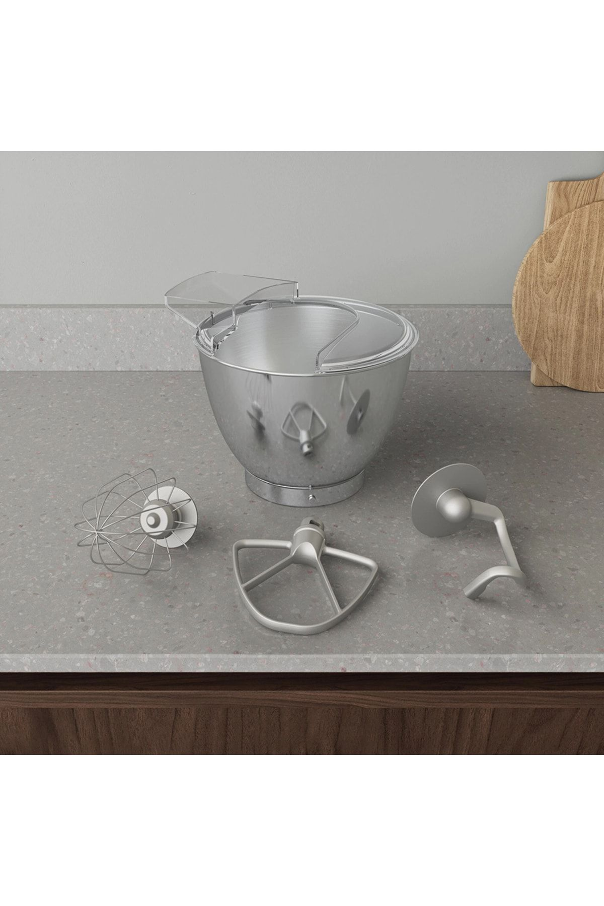 Una ciotola per mixer argentata con tre accessori, tra cui una frusta e un gancio per impastare, su un piano.