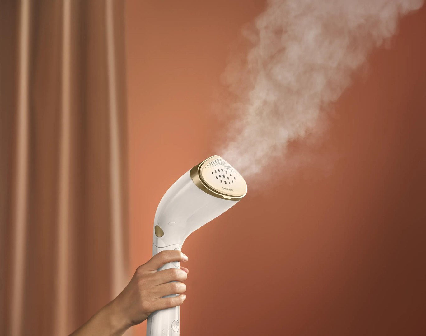 Una mano regge un vaporizzatore per indumenti bianco e oro che emette vapore su sfondo marrone.