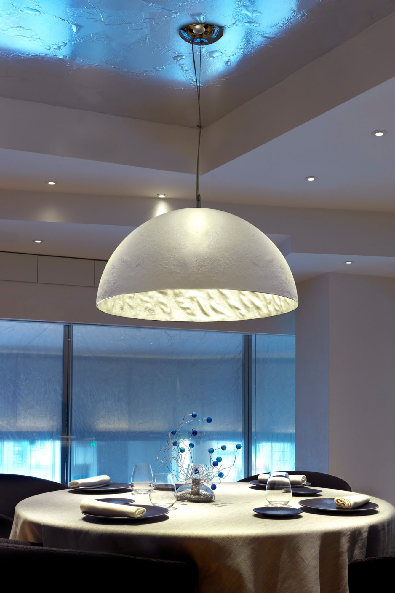 Un elegante tavolo da pranzo con decoro in vetro blu, coperti e una grande lampada semisferica sospesa sopra.