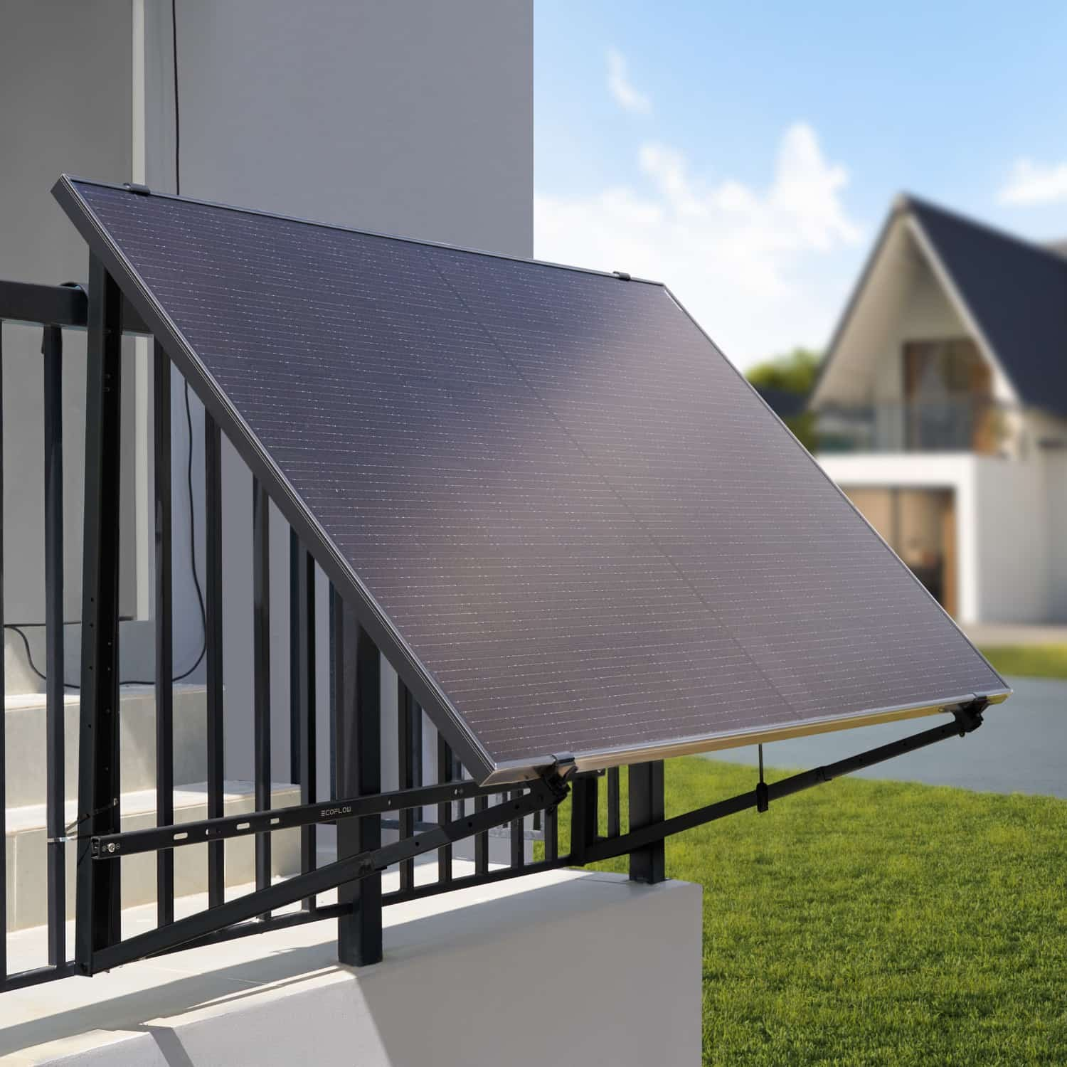 Un grande pannello solare è montato su una ringhiera nera, con una casa bianca e un prato verde sullo sfondo.