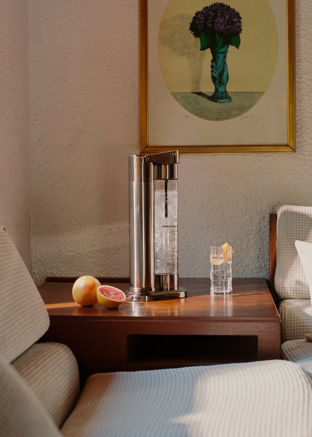 Una máquina de agua con gas plateada y pomelos en una mesa de madera, con un cuadro al fondo.