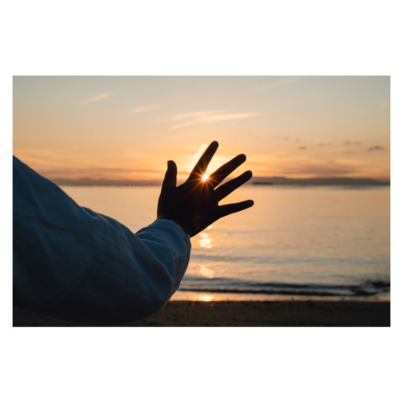 Die Hand einer Person greift nach einem Sonnenuntergang über dem Meer, die Sonne glimmt zwischen den Fingern.