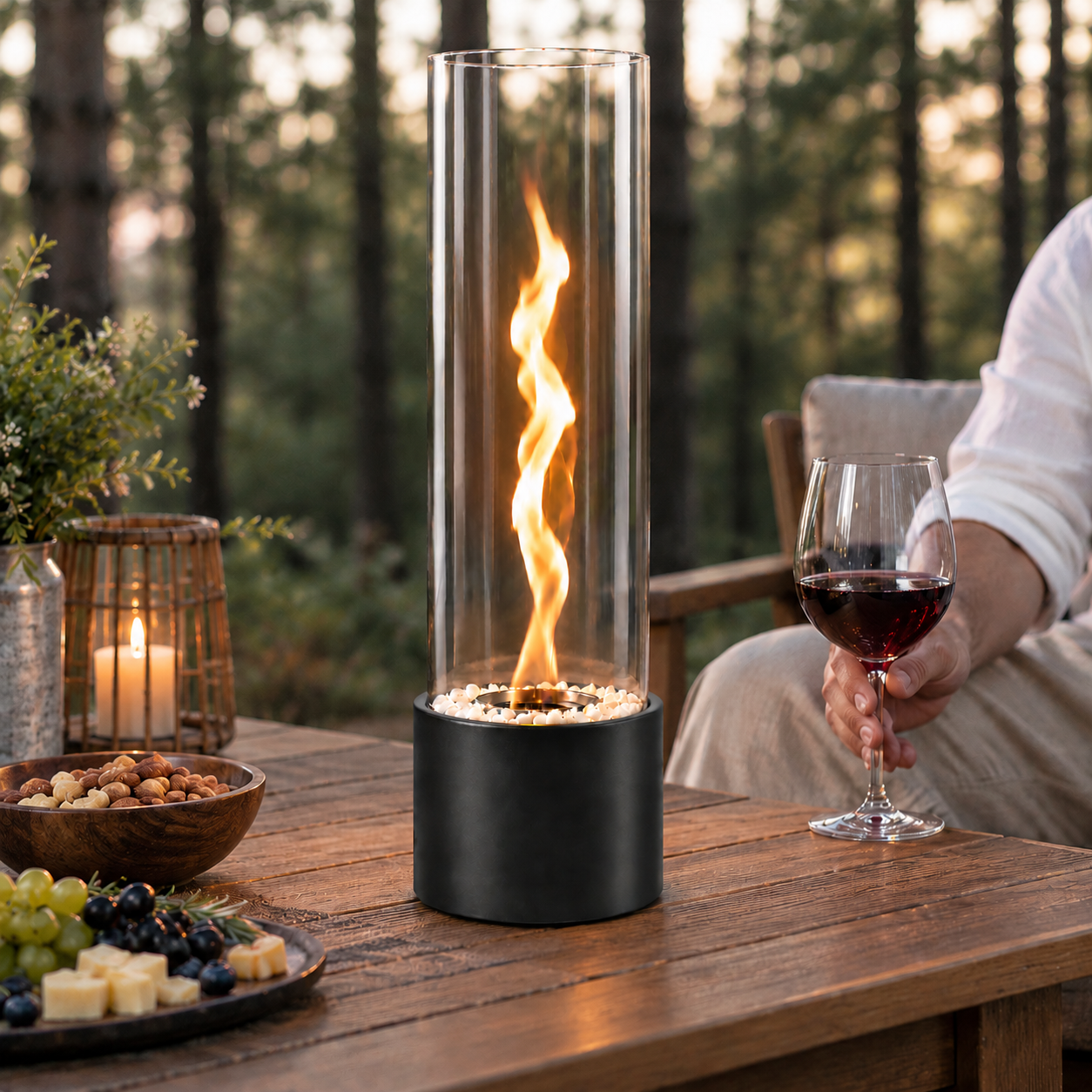 Ein gläserner Tischkamin mit Flamme brennt auf einem Holztisch mit Wein, Käse und Nüssen im Freien.
