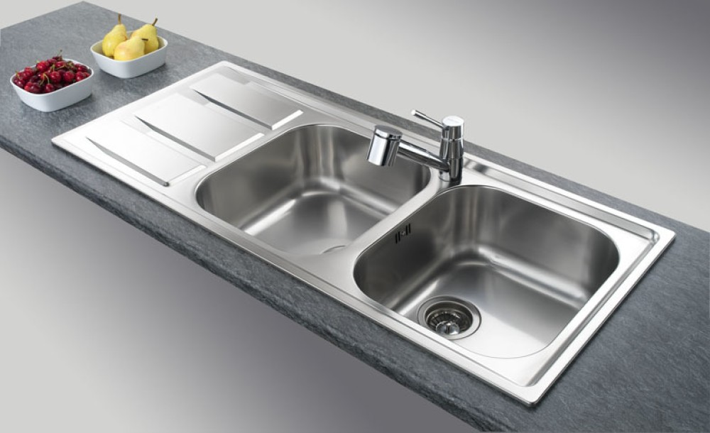 Lavello doppio in acciaio inox con rubinetto cromato su piano di lavoro scuro e ciotole di frutta.