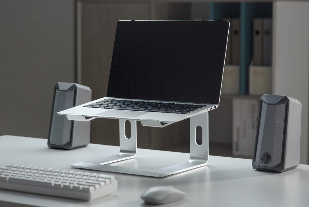 Ein Laptop auf einem Metallständer auf einem Schreibtisch, flanked von zwei grauen Lautsprechern und einer Tastatur davor.