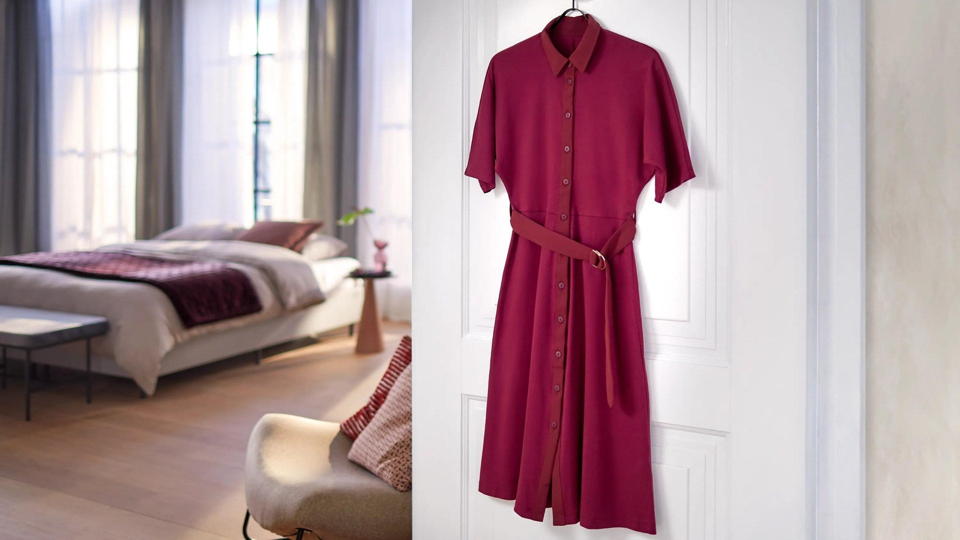 Un abito camicia bordeaux con cintura è appeso a una porta bianca, con una camera da letto sullo sfondo.