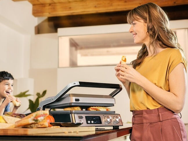 Una donna con una maglietta gialla mangia un panino accanto a una piastra, mentre un bambino sorride sullo sfondo.