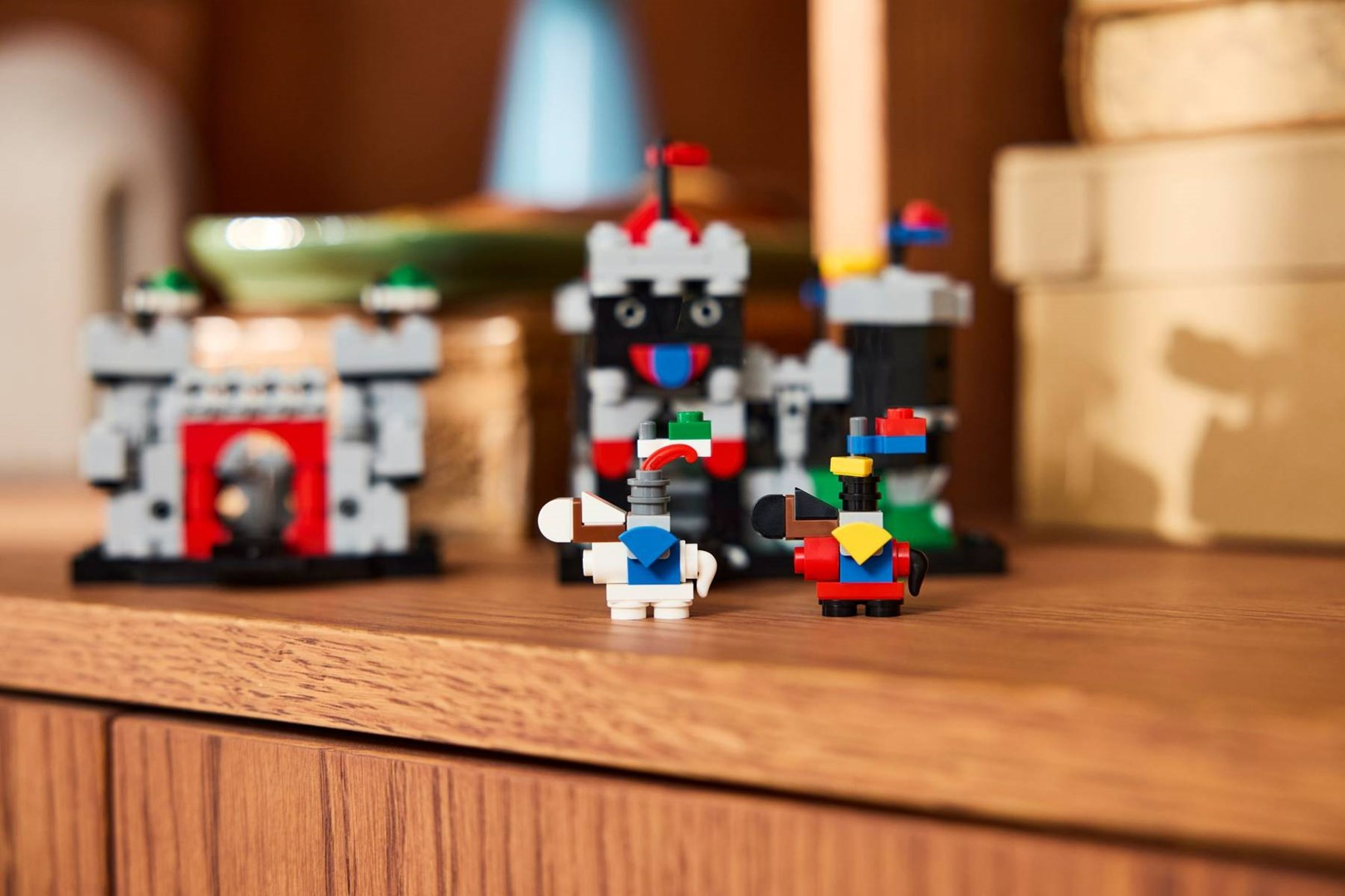 Ahşap bir rafta iki Lego kalesi ve iki şövalye figürü, arkada bulanık nesneler var.
