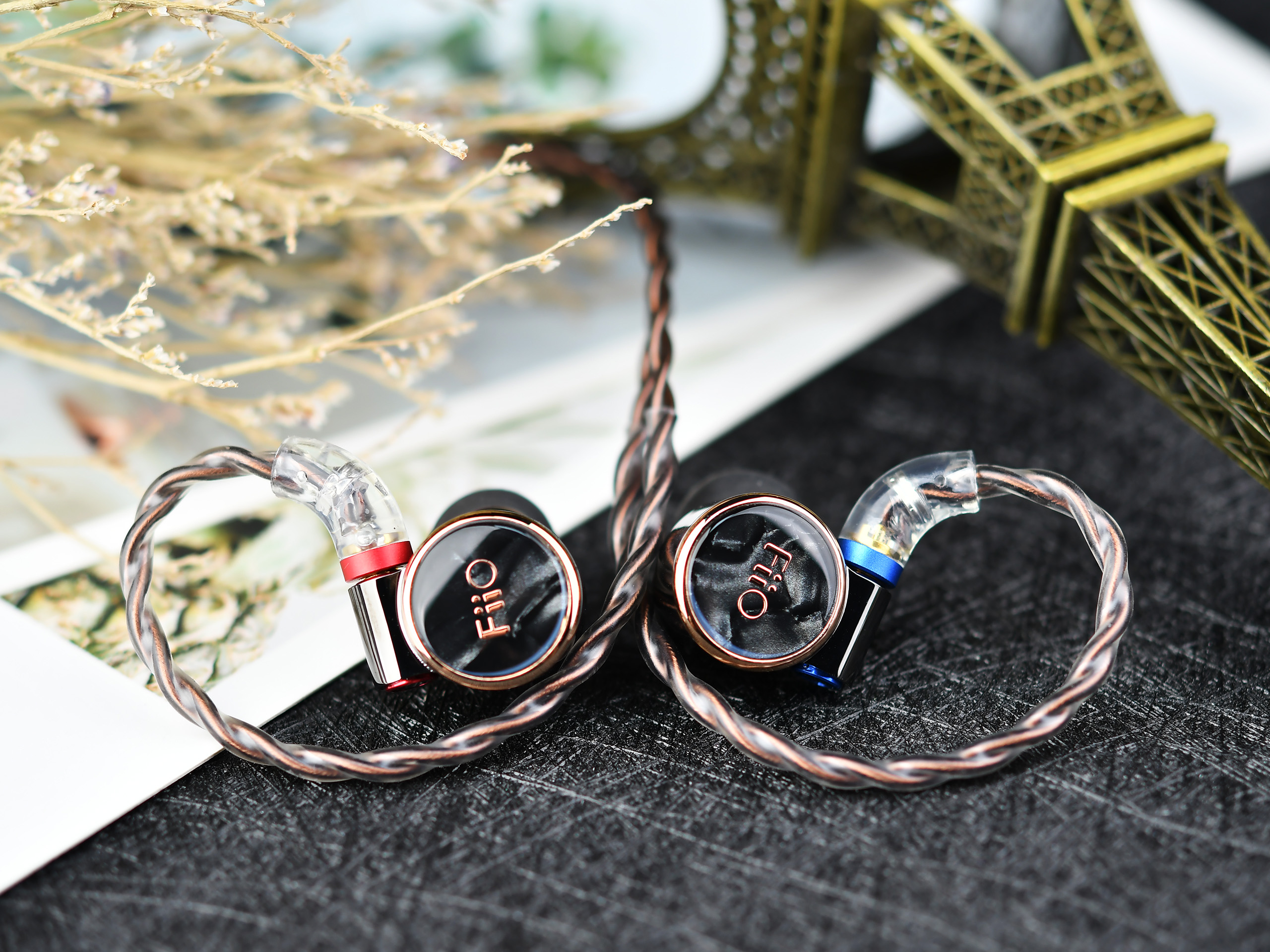 Auriculares intraurales con cable de cobre sobre superficie negra, con flores secas y miniatura de la Torre Eiffel al fondo.