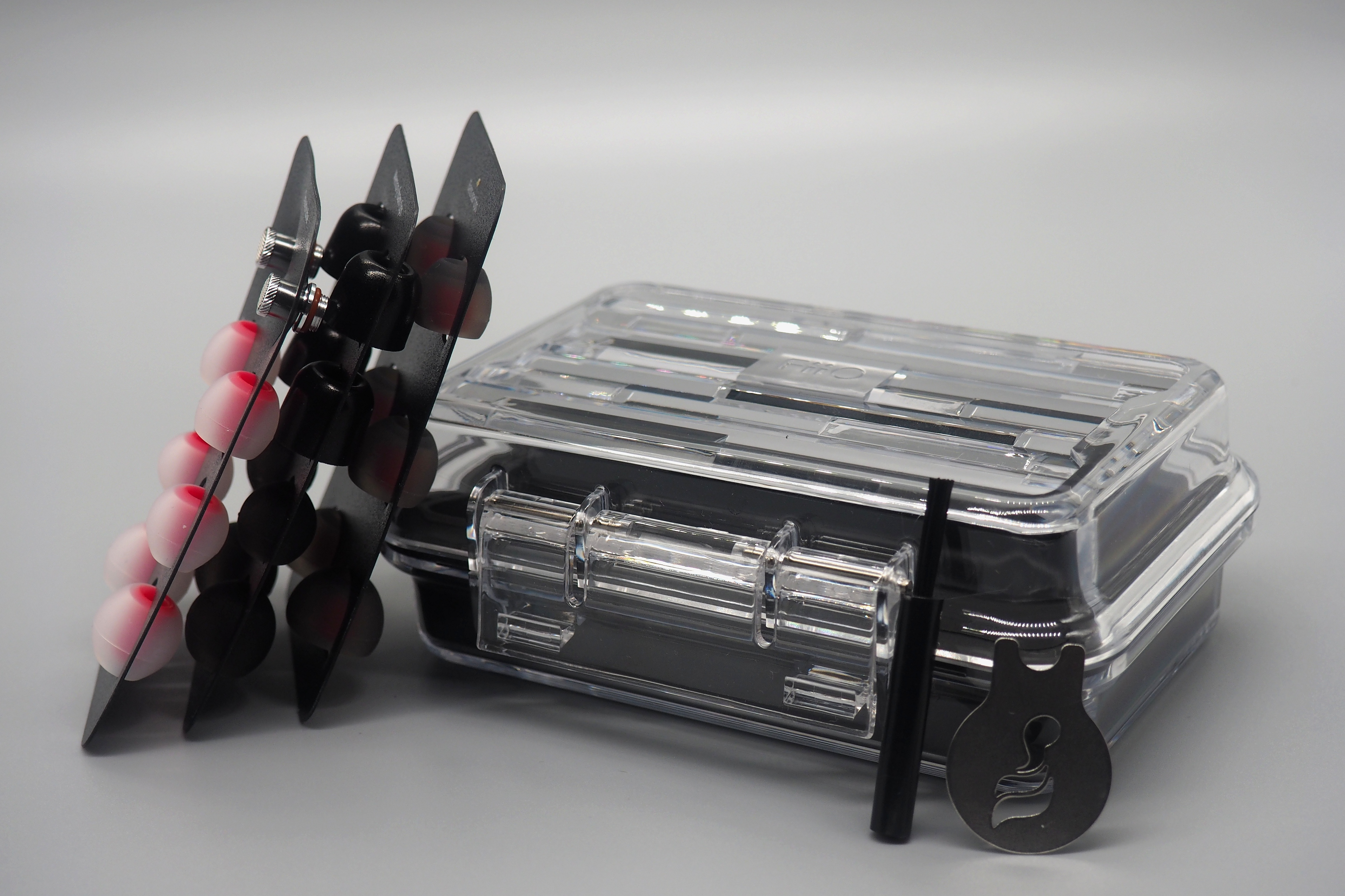 Caja transparente, soportes negros con almohadillas, pincel pequeño y herramienta de metal sobre fondo gris.