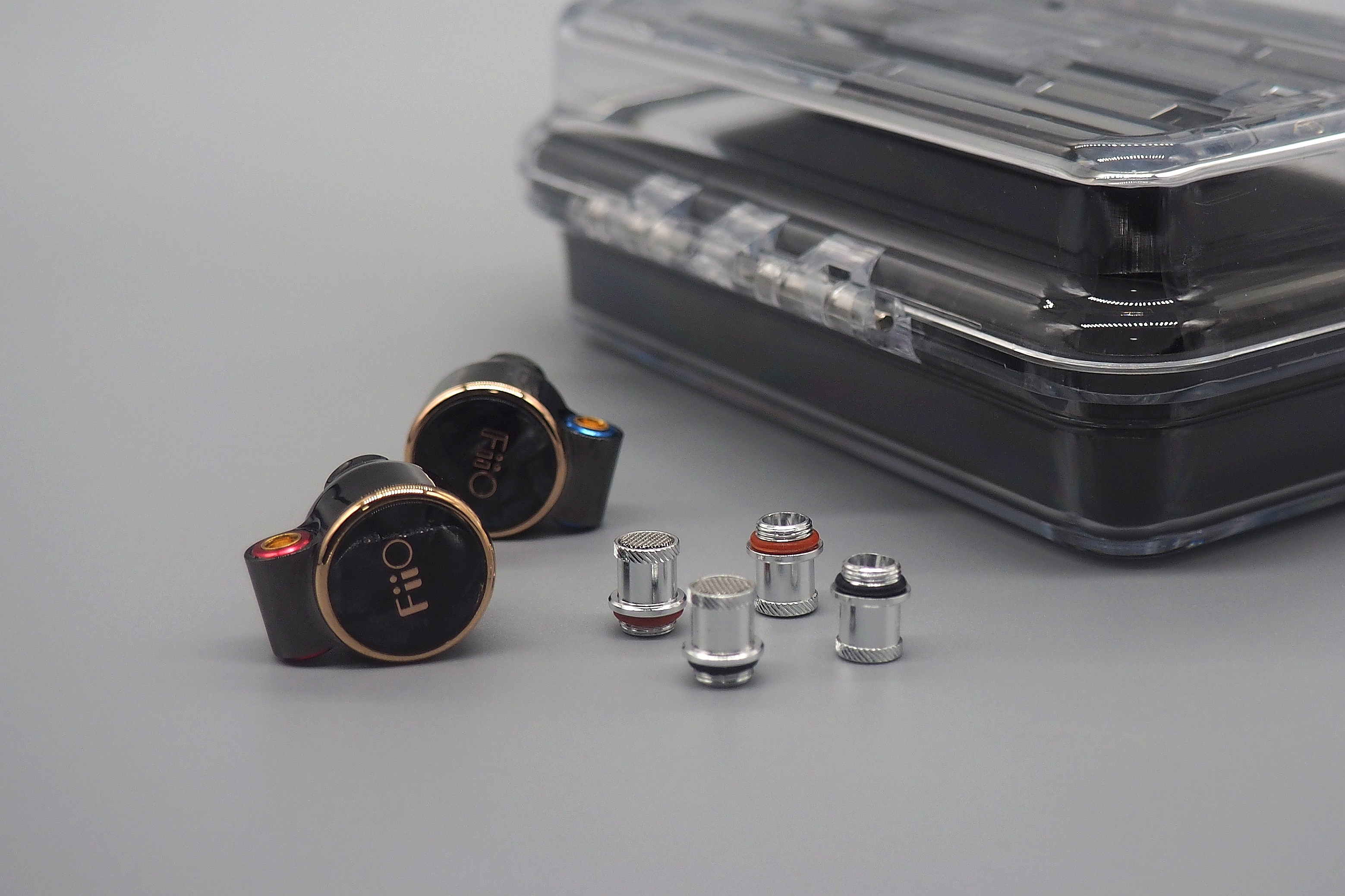 Dos auriculares negros FiiO y cuatro filtros metálicos frente a un estuche transparente sobre fondo gris.