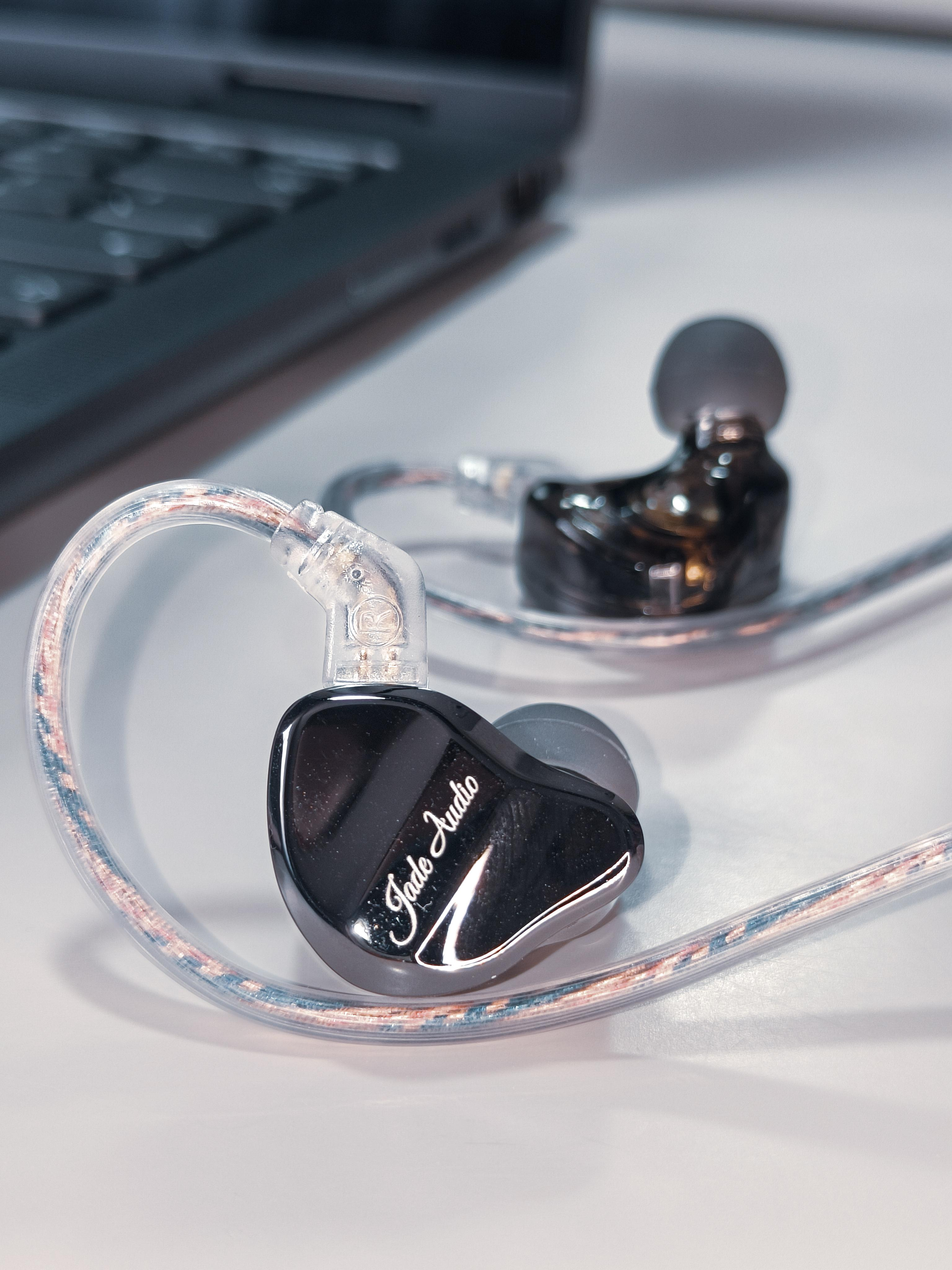 Przewodowe słuchawki dokanałowe Jade Audio z przezroczystymi kablami na białym biurku obok laptopa.