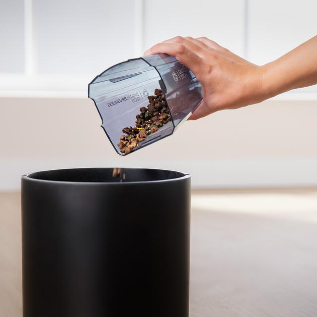Una mano vierte residuos de un depósito de aspiradora en un cubo negro.