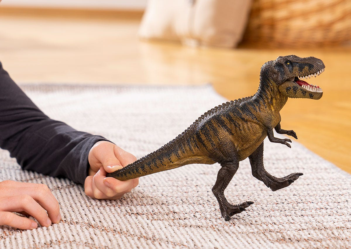Ein Kind hält den Schwanz einer realistischen Tyrannosaurus-Rex-Spielzeugfigur auf einem Teppich.