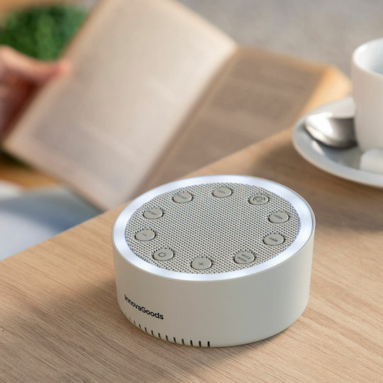 Una máquina de sonido redonda blanca con botones está sobre una mesa de madera, con un libro abierto y una taza de café.