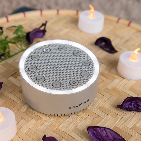 Una máquina de sonido blanca y redonda con botones, rodeada de velas y pétalos morados en una bandeja de bambú.