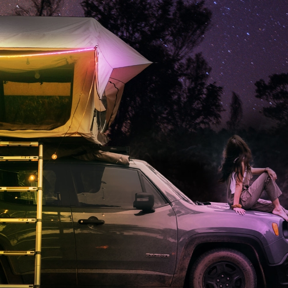 Una persona está sentada en el capó de un coche bajo una tienda de techo y un cielo estrellado.
