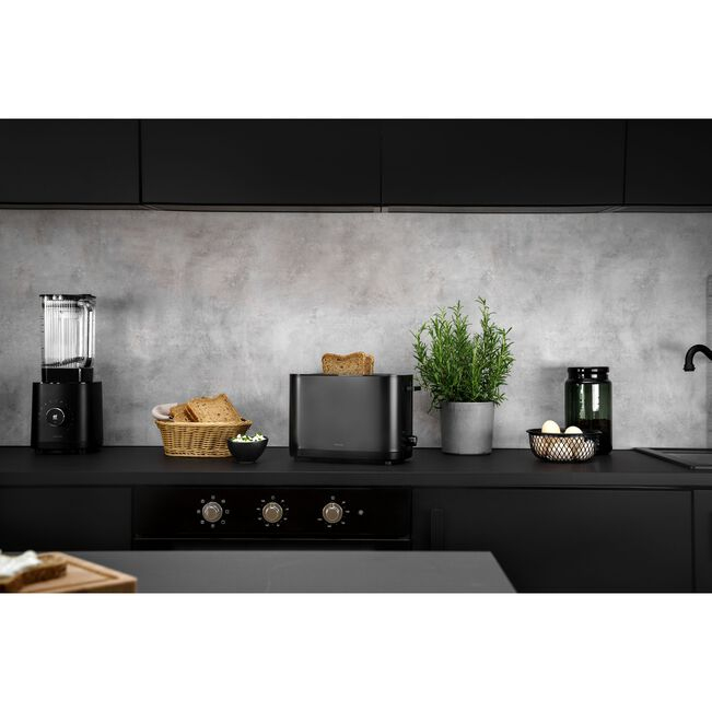 Cocina moderna con tostadora negra, licuadora, panera y planta verde en encimera oscura.