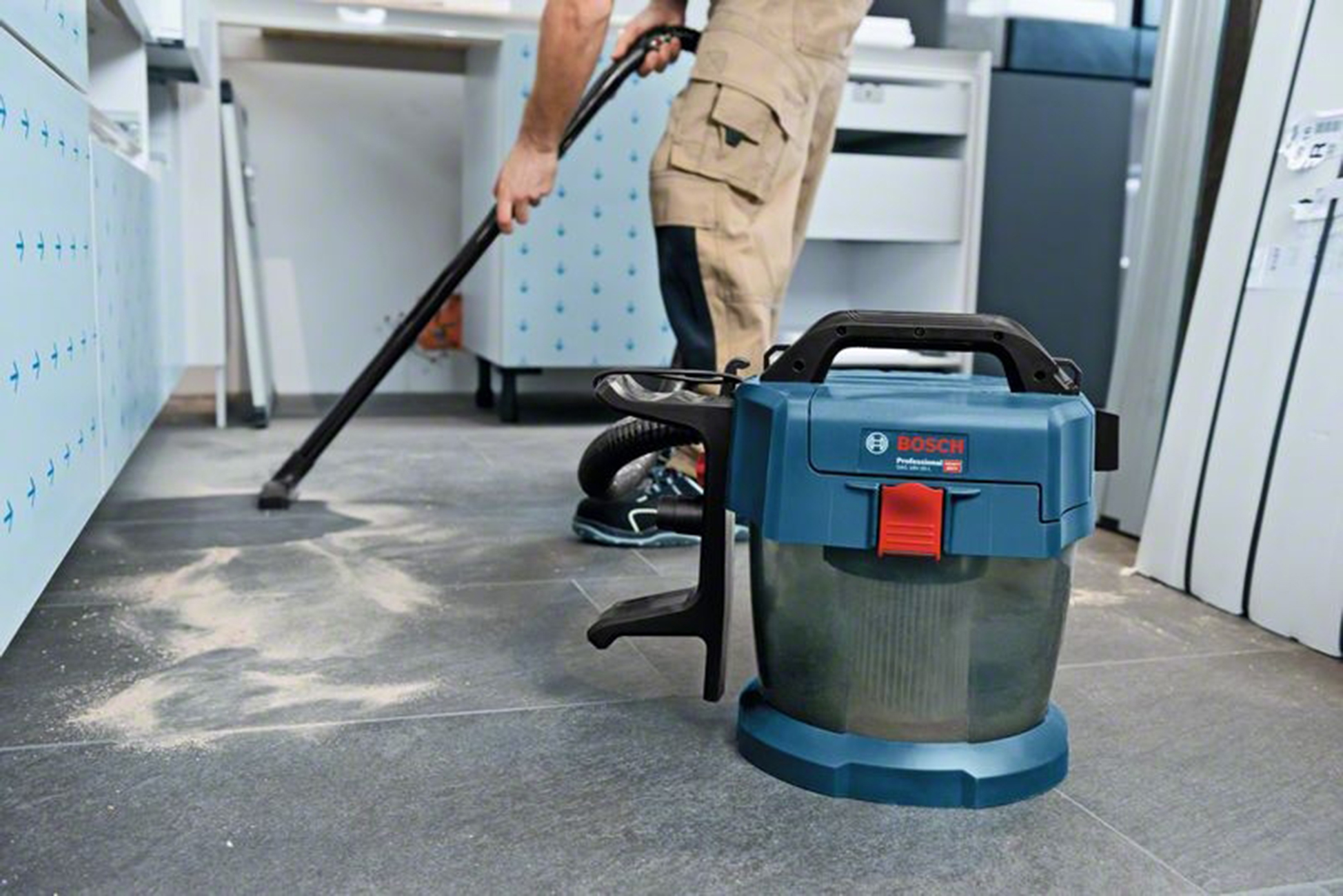 Bir adam bitmemiş bir odada mavi bir Bosch elektrikli süpürgeyle fayans zemini temizliyor.