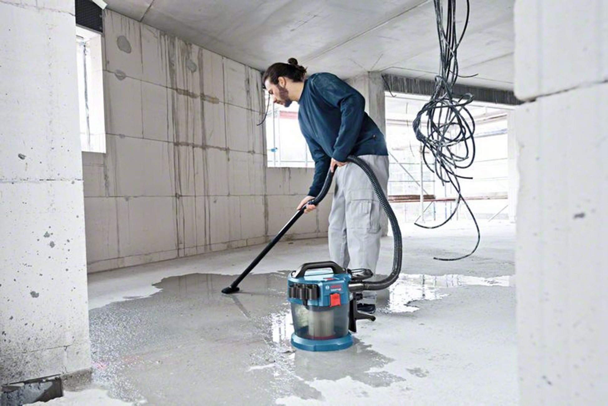 Yarım kalmış bir odada bir adam beton zemindeki suyu vakumluyor.