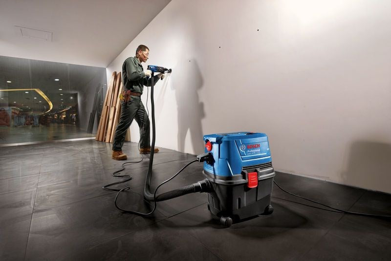 Un uomo in abiti da lavoro verdi fora una parete bianca, collegato a un aspirapolvere blu Bosch.