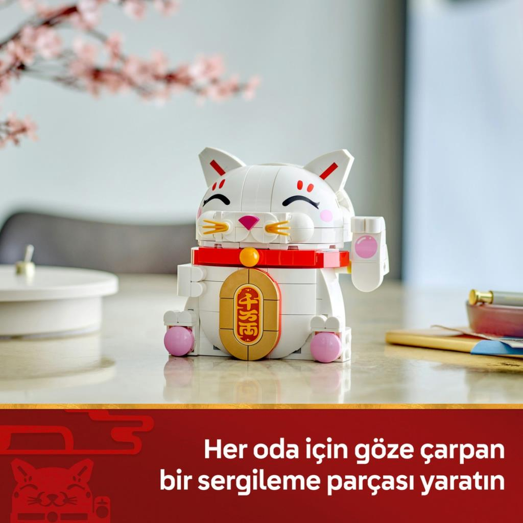 Ahşap masa üzerinde beyaz bir LEGO şans kedisi, arka planda pembe çiçekler var.