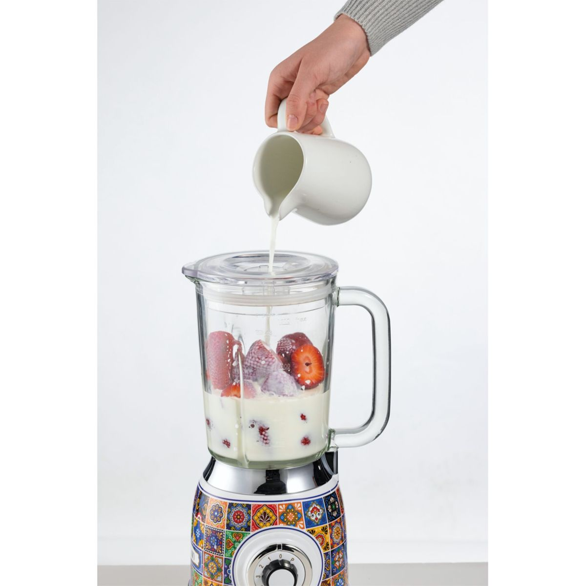 Bir el, çilek ve ahududu dolu bir blender'a küçük bir sürahiden süt döküyor.