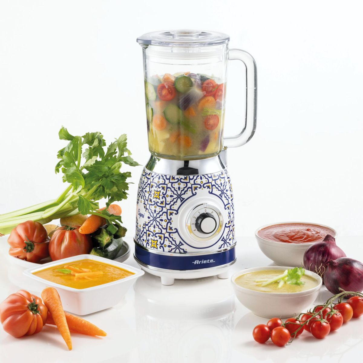 Sebzelerle dolu desenli mavi bir blender, taze domatesler, havuçlar ve çorba kaseleriyle çevrili.