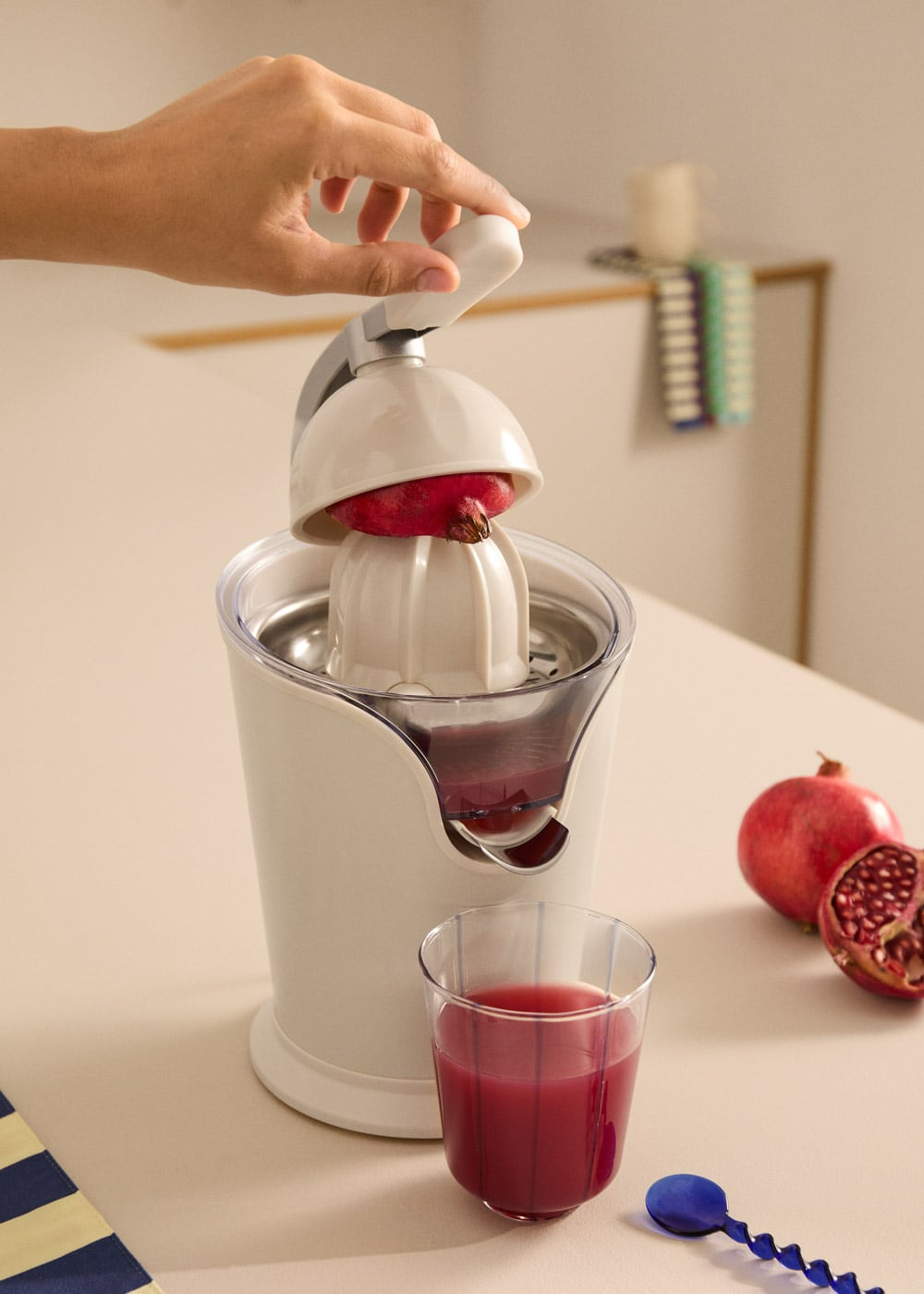 Una mano presiona un exprimidor blanco con medio granada, junto a un vaso de jugo rojo y granadas en una mesa.