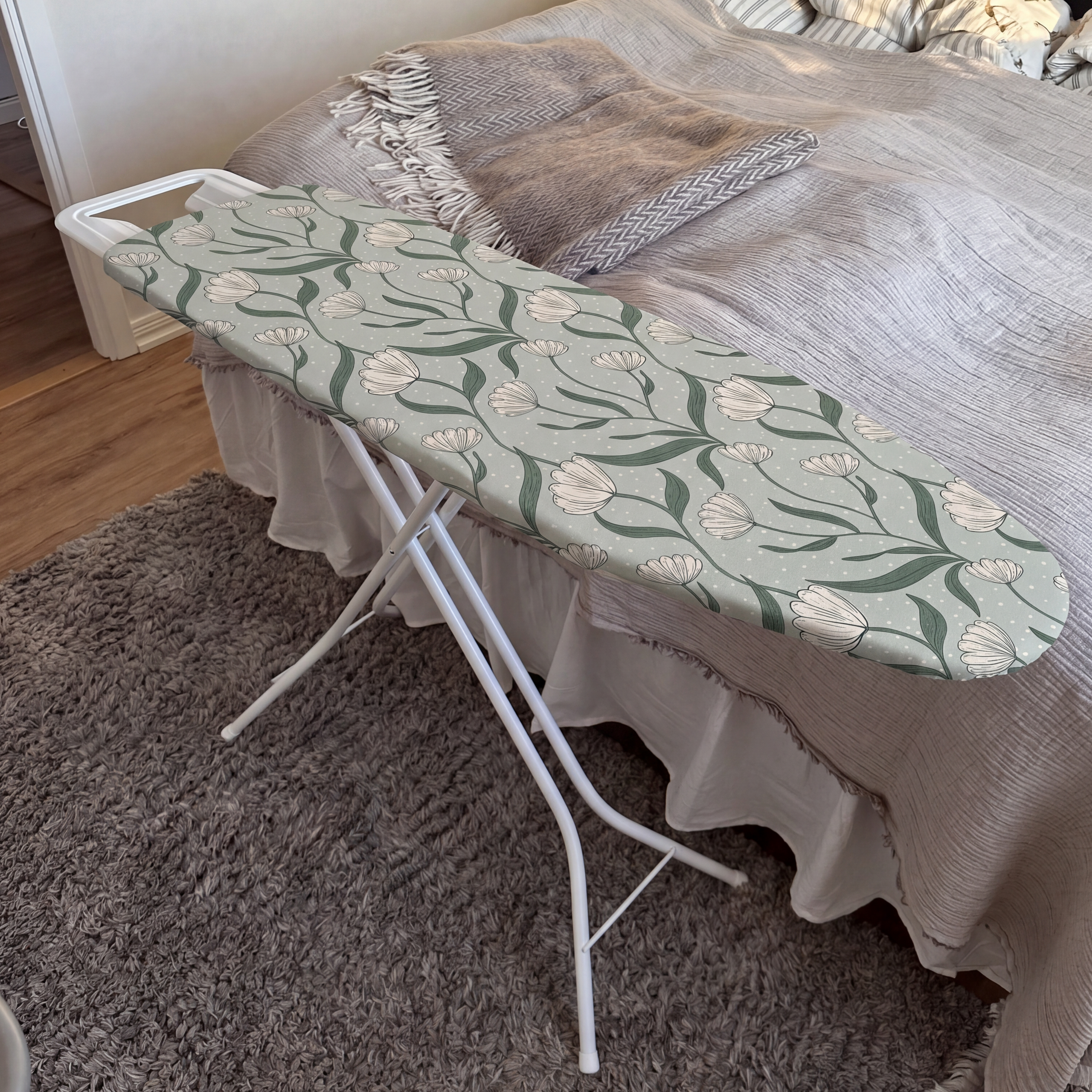 Una tabla de planchar con funda floral en un dormitorio sobre una alfombra gris, con una cama hecha al fondo.
