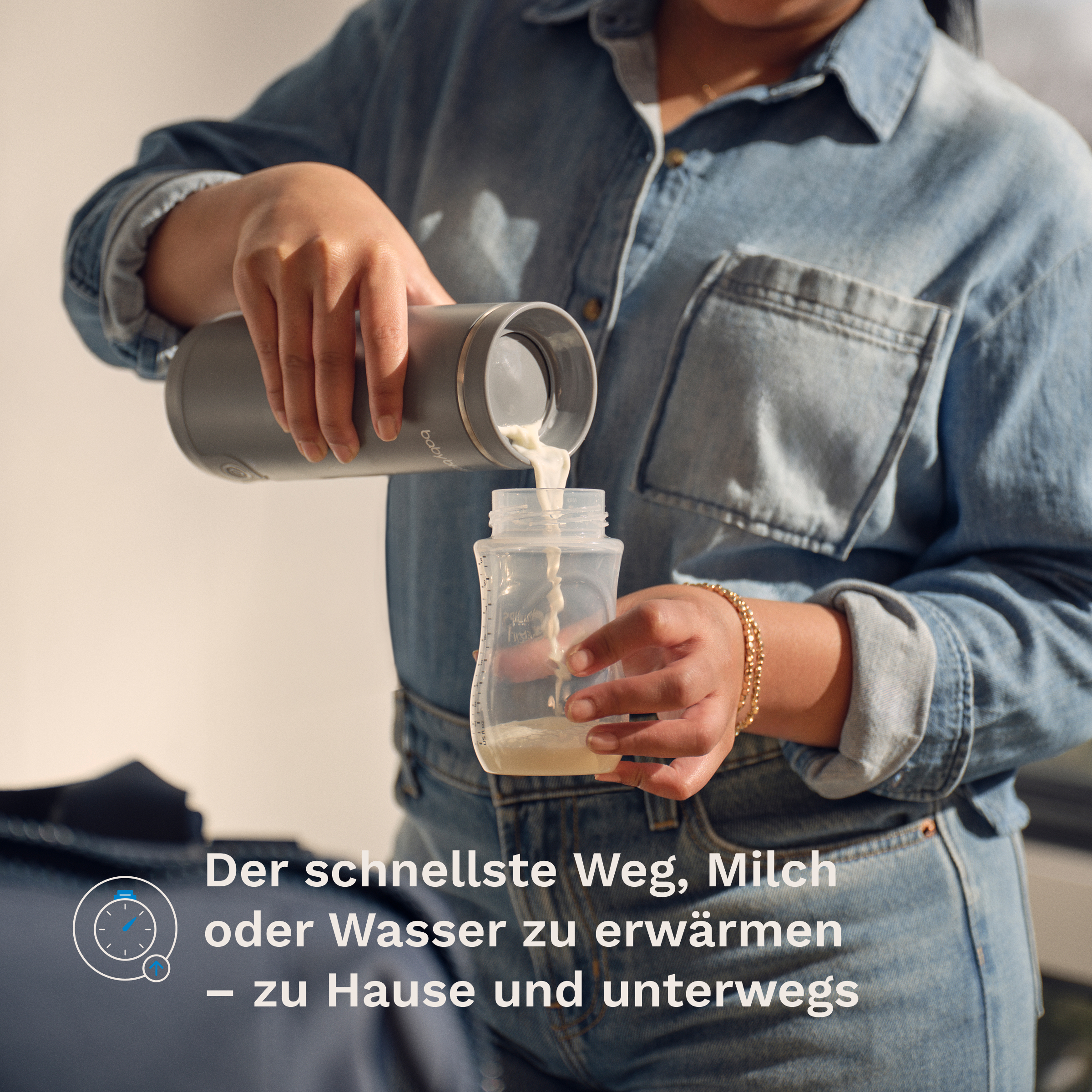 Eine Person im Jeanshemd gießt Milch aus einer grauen Thermoskanne in ein Babyfläschchen.
