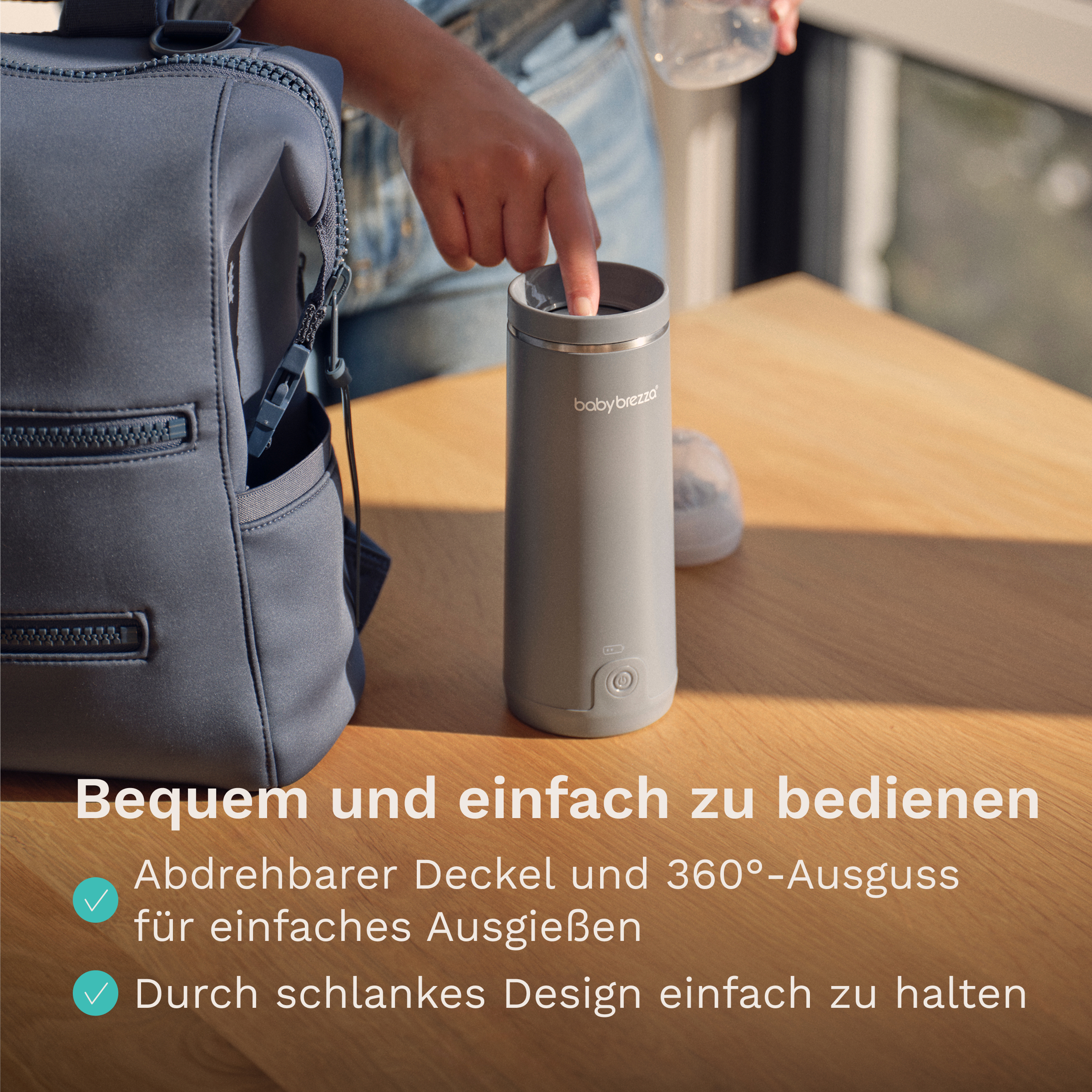 Eine Hand bedient einen grauen Baby Brezza Flaschenwärmer auf einem Holztisch neben einem grauen Rucksack.