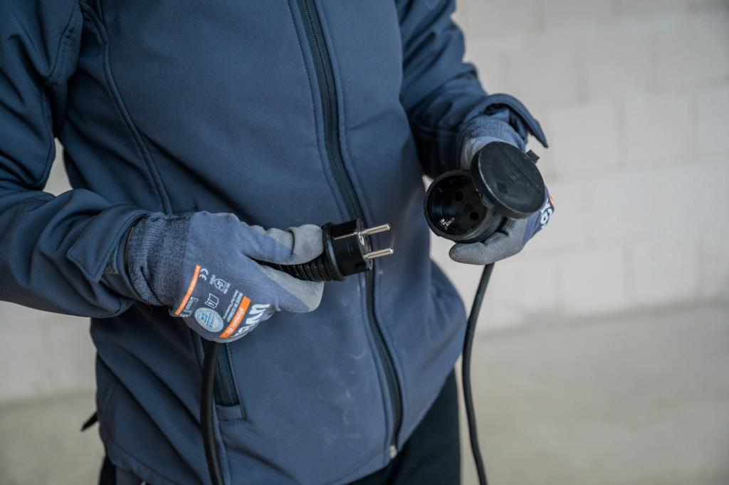 Eine Person in blauer Jacke und Arbeitshandschuhen hält einen schwarzen Stecker und eine Steckdose.