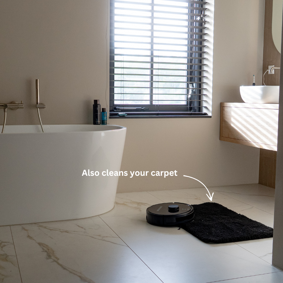 Ein Saugroboter reinigt einen kleinen schwarzen Badteppich auf weißem Fliesenboden neben einer Badewanne.