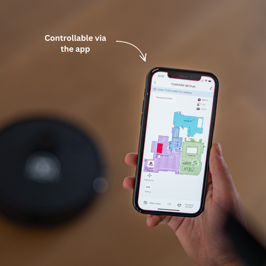 Eine Hand hält ein Smartphone mit einer Roboterstaubsauger-App, während das Gerät im Hintergrund auf einem Holzboden steht.