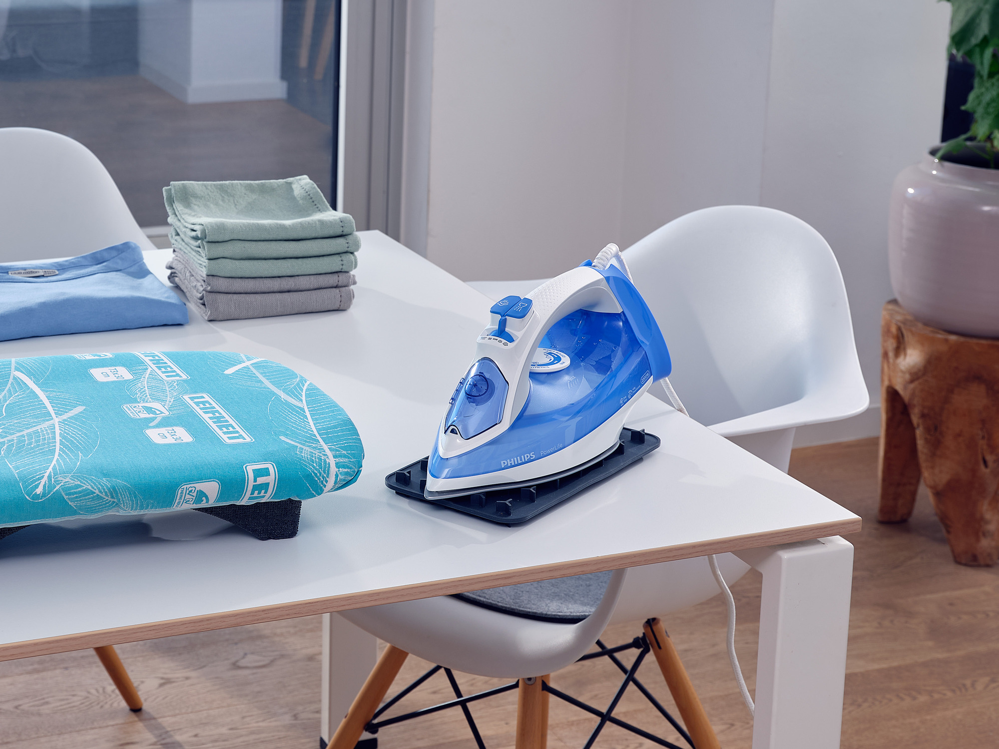 Blau-weiße Philips Bügeleisen auf einem weißen Tisch mit gefalteter Kleidung und Bügelbrett.